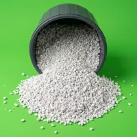 White granular pellets spilling from a black bucket onto a bright green surface, forming a mound on the ground.