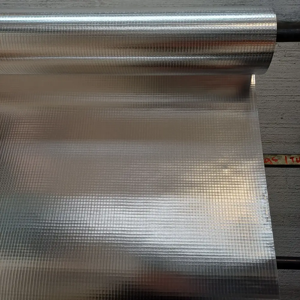 Roll of shiny textured aluminum/foil-like material partially unrolled on a wooden surface, showing a grid pattern on the sheet.